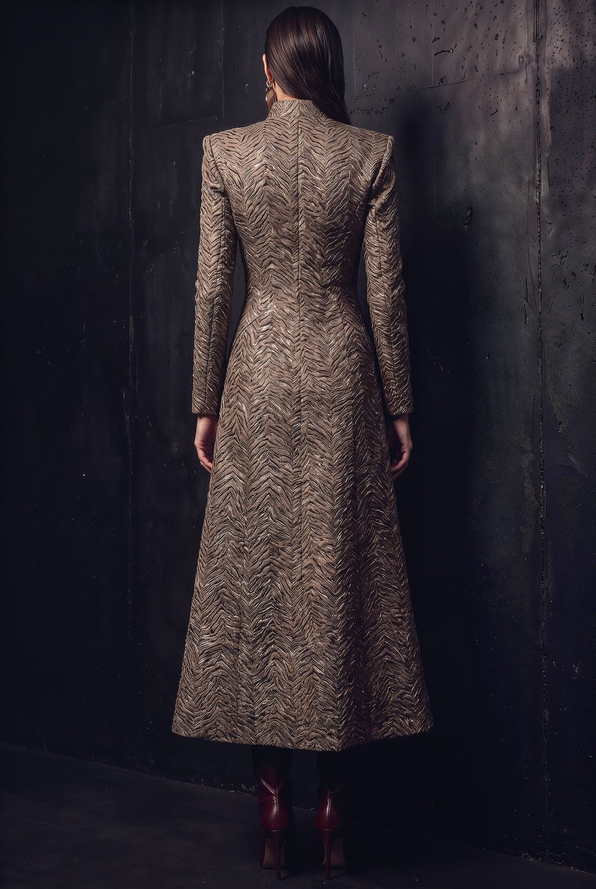 A woman viewed from behind wearing a long-sleeved, high-neck midi coat with a textured chevron pattern in brown and beige, standing against a dark textured wall.
