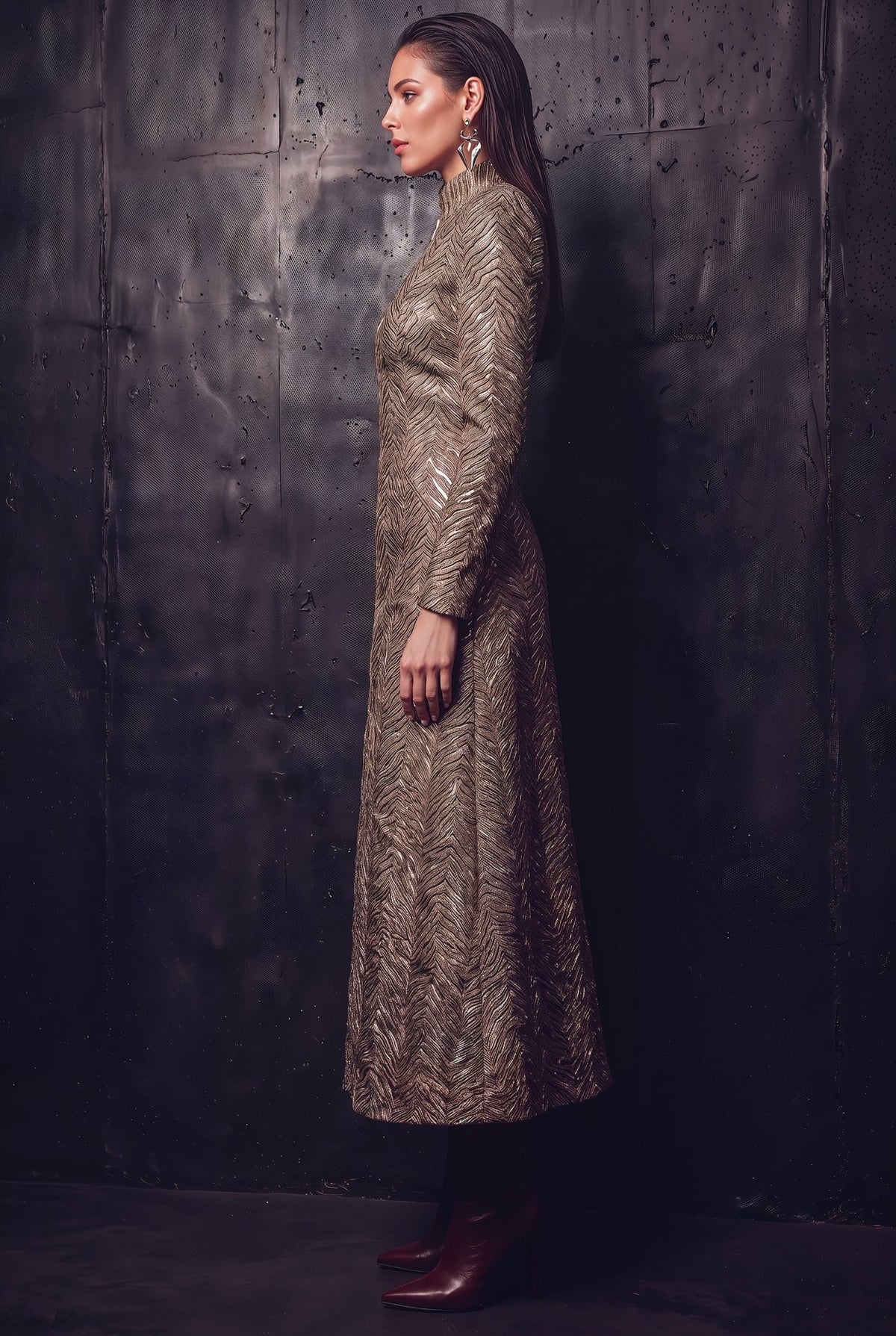 A woman stands in profile wearing a long-sleeved, high-neck maxi dress with an intricate metallic gold and brown abstract pattern, paired with dark brown leather boots, set against a dark textured wall.