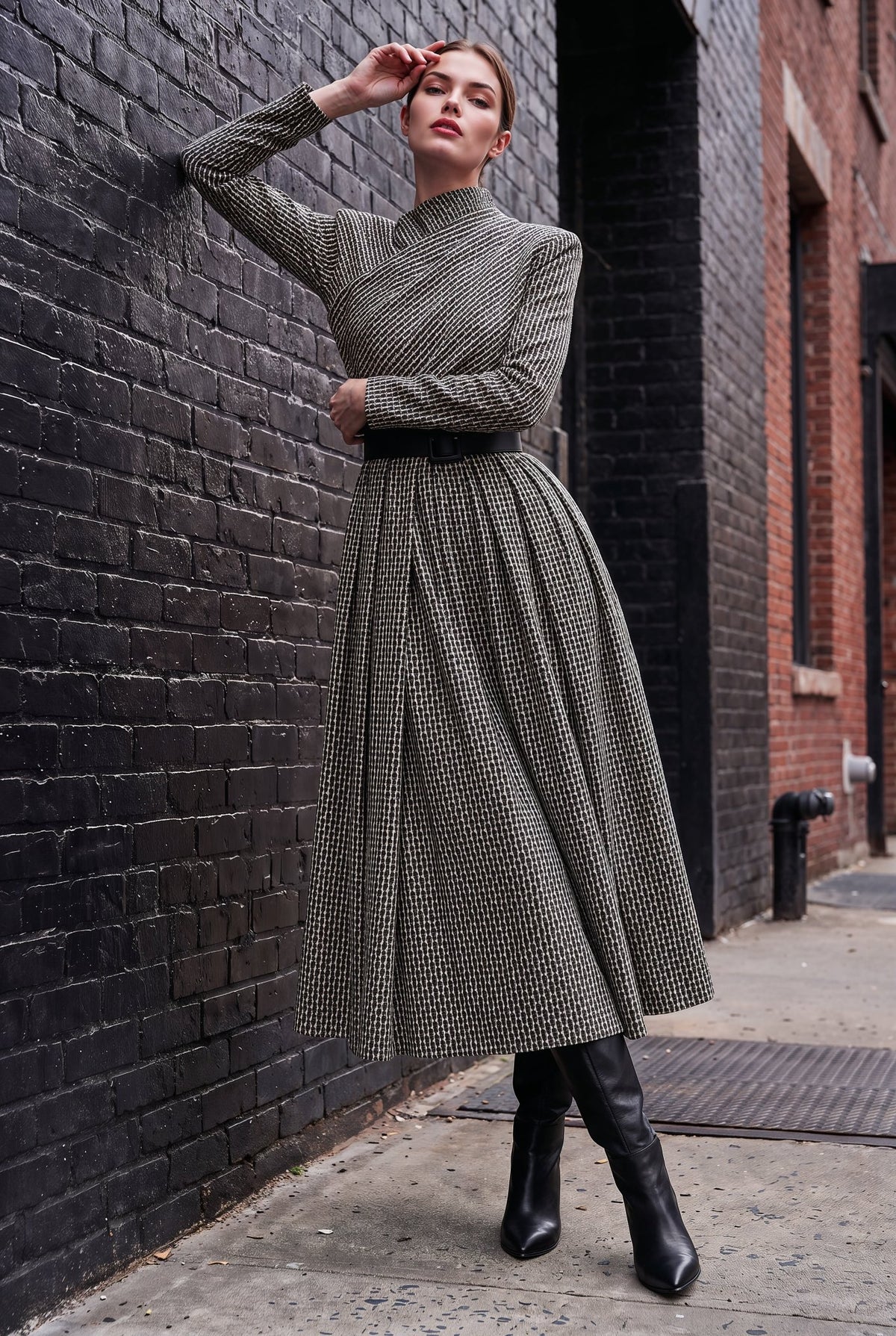 A woman models a long-sleeve, high-neck midi dress with a textured black and beige houndstooth pattern, styled with a black belt and black knee-high boots, posing against a dark brick wall.