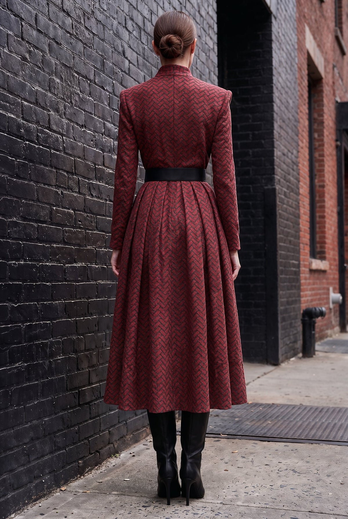 A woman viewed from behind wearing a long-sleeved, high-neck midi dress with a textured chevron pattern in deep red and black, featuring a black belt at the waist. She is standing on a city sidewalk between a black brick wall and a red brick building.