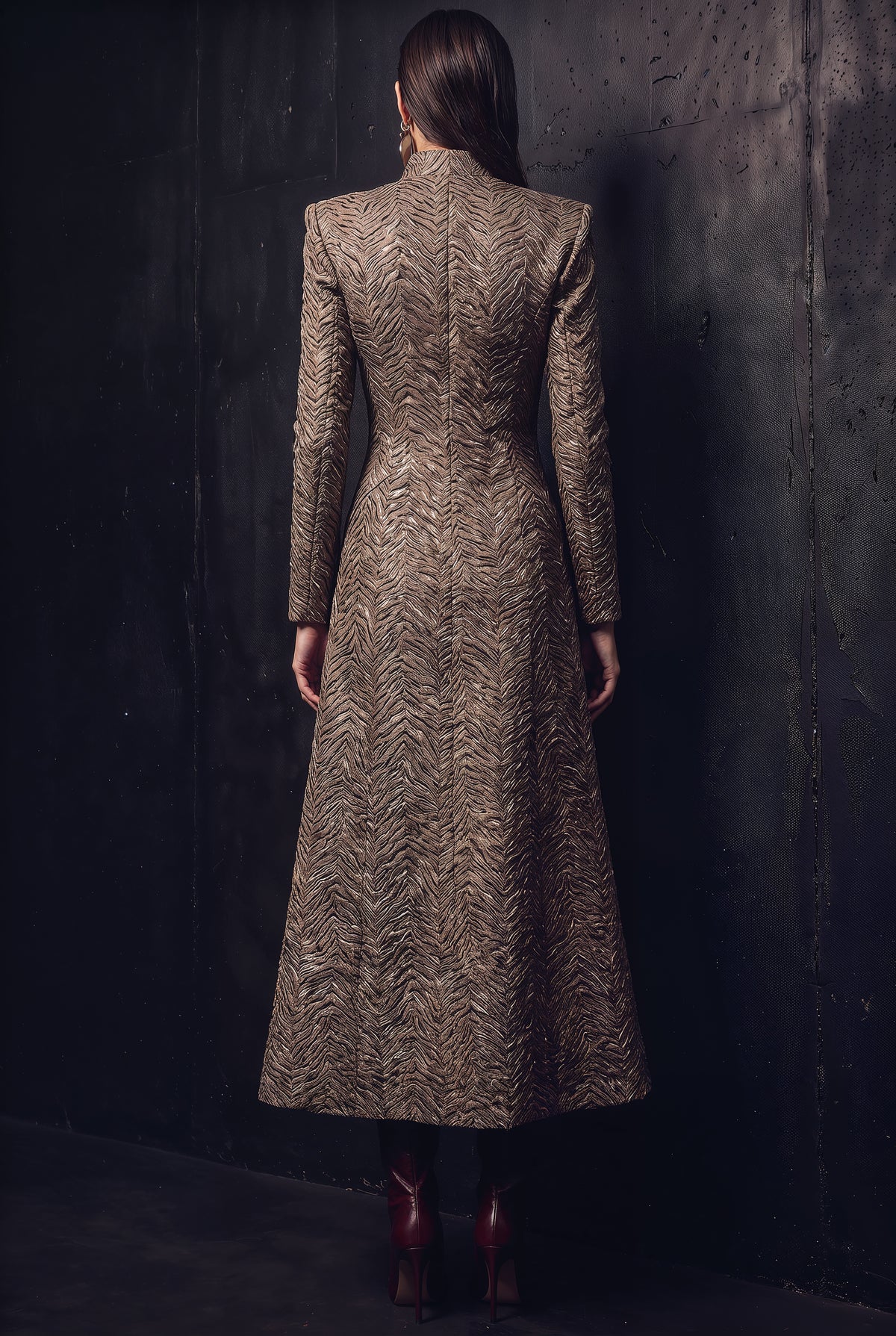 A woman viewed from behind wearing a long-sleeved, high-neck midi coat with a textured chevron pattern in brown and beige, standing against a dark textured wall.