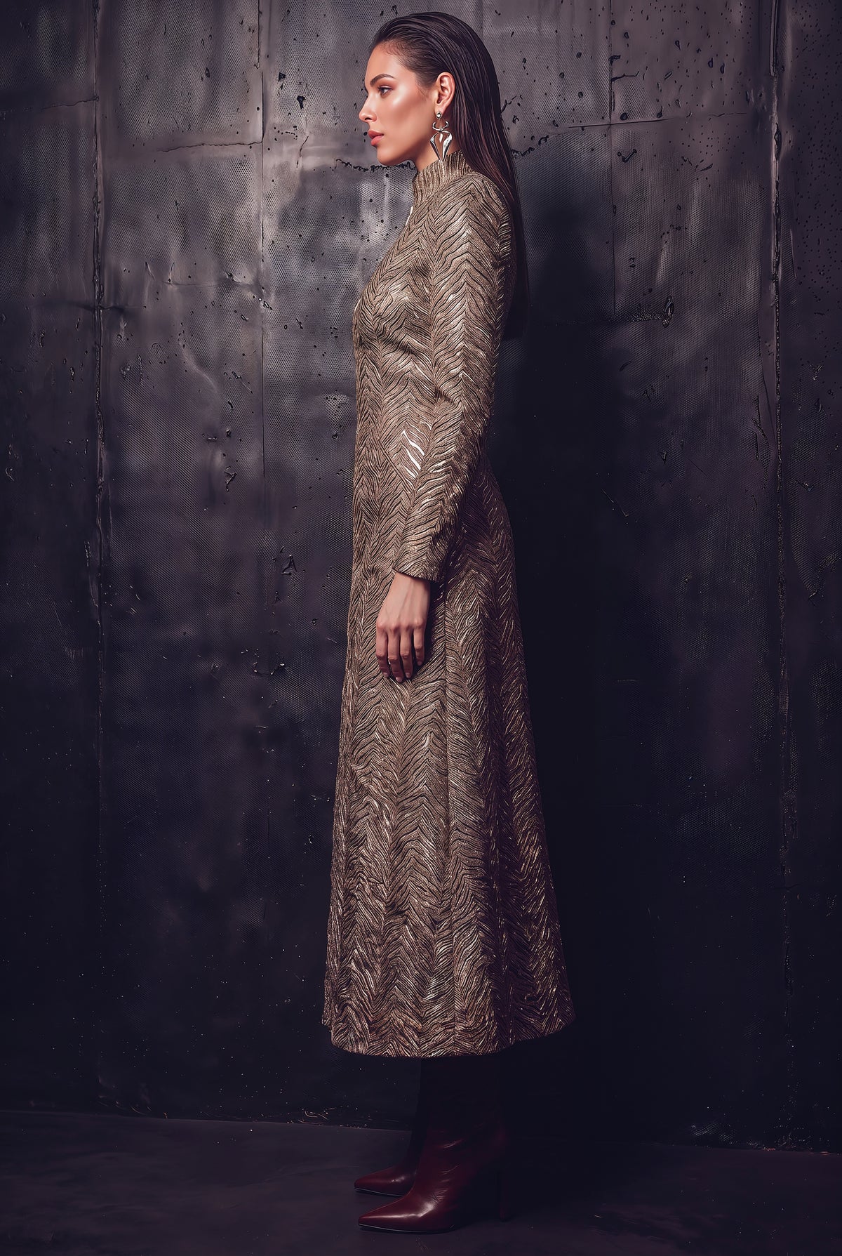 A woman stands in profile wearing a long-sleeved, high-neck maxi dress with an intricate metallic gold and brown abstract pattern, paired with dark brown leather boots, set against a dark textured wall.