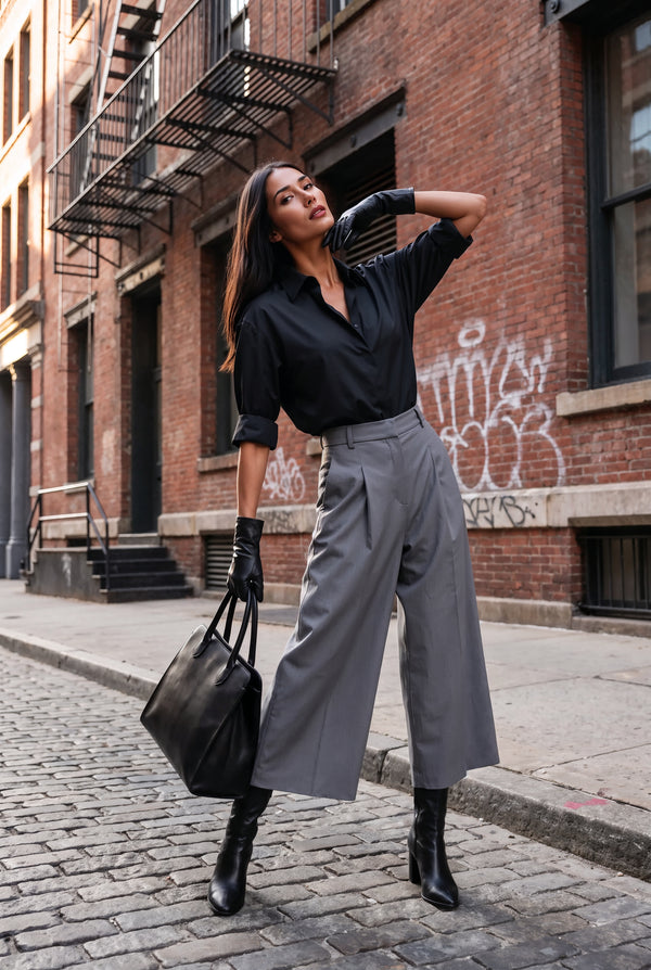 A woman stands on a city street wearing a black button-up shirt, gray wide-leg cropped pants, black gloves, and black ankle boots, while carrying a large black tote bag. Brick buildings with fire escapes and graffiti are in the background.
