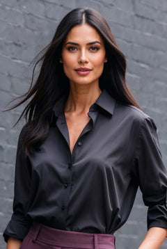 A woman with long dark hair wearing a black satin button-down shirt and purple trousers, standing against a gray brick wall.