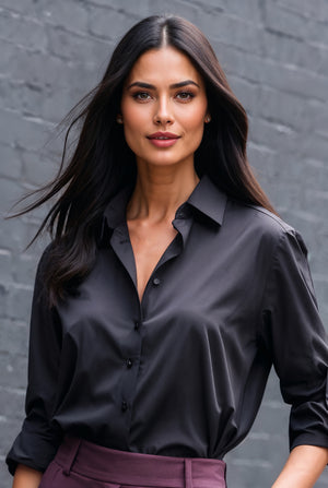 A woman with long dark hair wearing a black satin button-down shirt and purple trousers, standing against a gray brick wall.