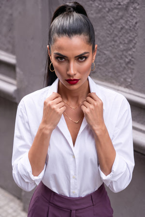 A woman with her hair in a high ponytail wearing a white button-down shirt and purple trousers, posing against a gray textured wall.