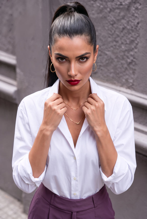 A woman with her hair in a high ponytail wearing a white button-down shirt and purple trousers, posing against a gray textured wall.