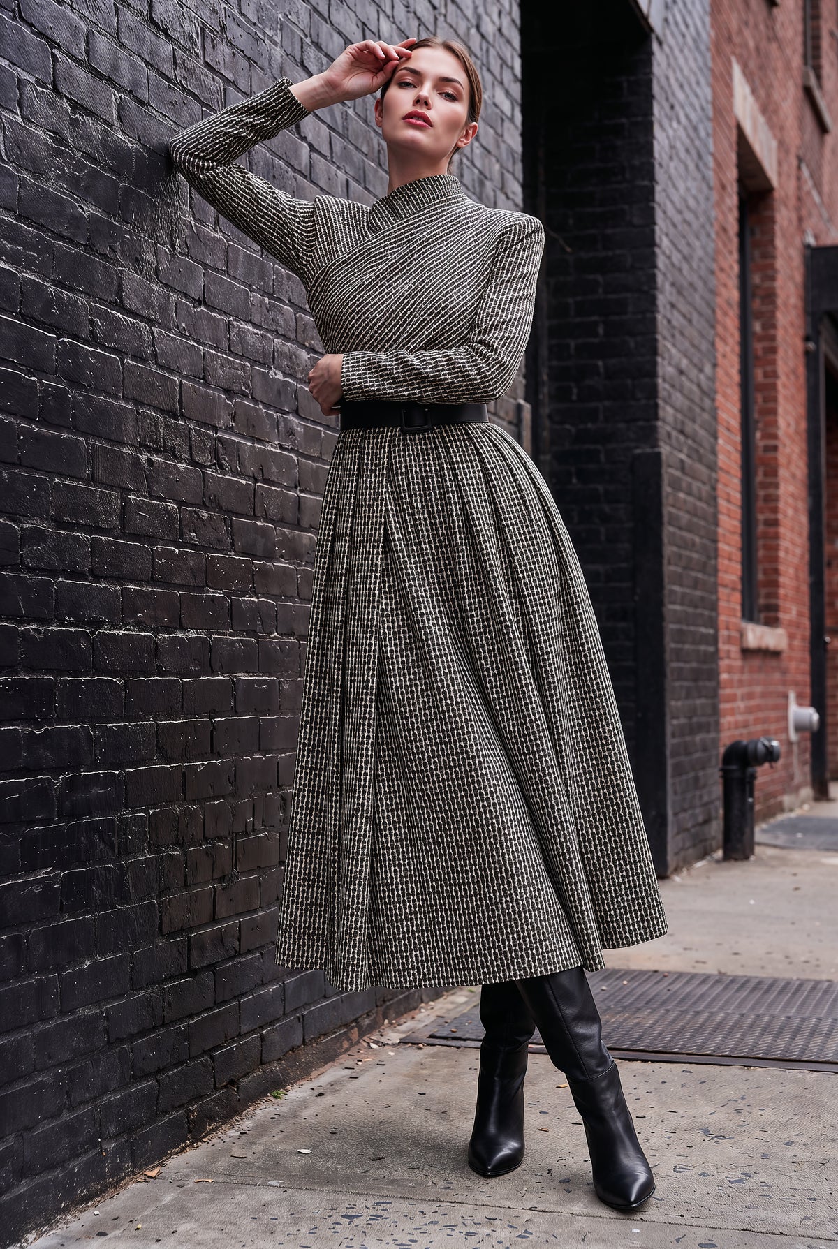A woman models a long-sleeve, high-neck midi dress with a textured black and beige houndstooth pattern, styled with a black belt and black knee-high boots, posing against a dark brick wall.