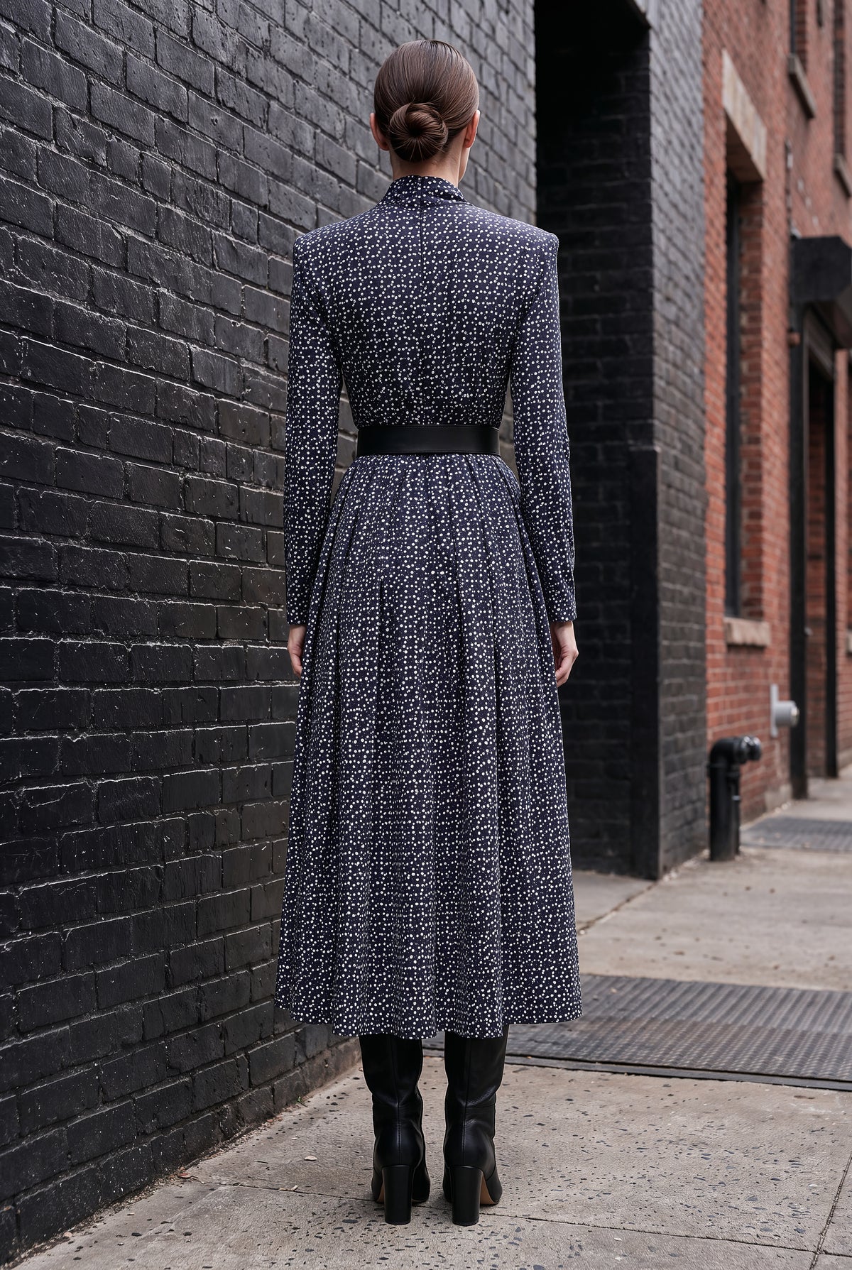 A woman viewed from behind wearing a long-sleeved midi dress with a textured black and white geometric pattern, a black waist belt, and black ankle boots, standing on a city sidewalk next to a black brick wall.