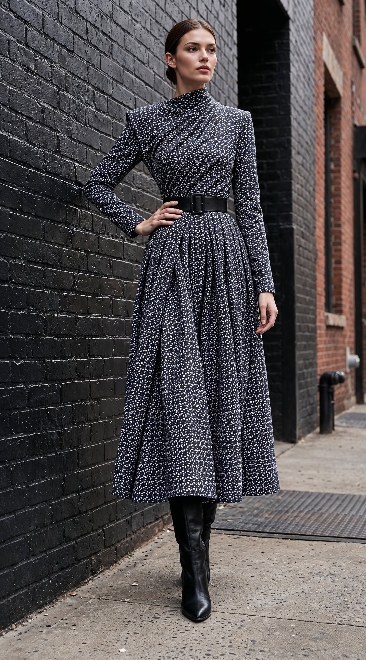 A woman stands on a city sidewalk against a black brick wall, wearing a long-sleeve high-neck midi dress with a black and white geometric pattern, a black belt, and black ankle boots.