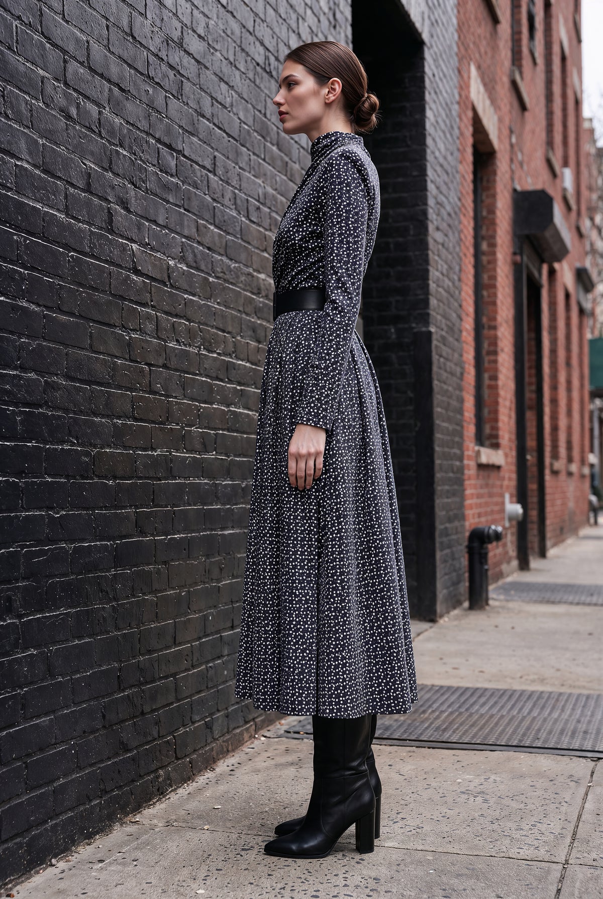 A woman stands in profile on a city sidewalk beside a dark brick wall, wearing a long-sleeve midi dress with a black and gray geometric pattern, a black belt, and black heeled ankle boots.