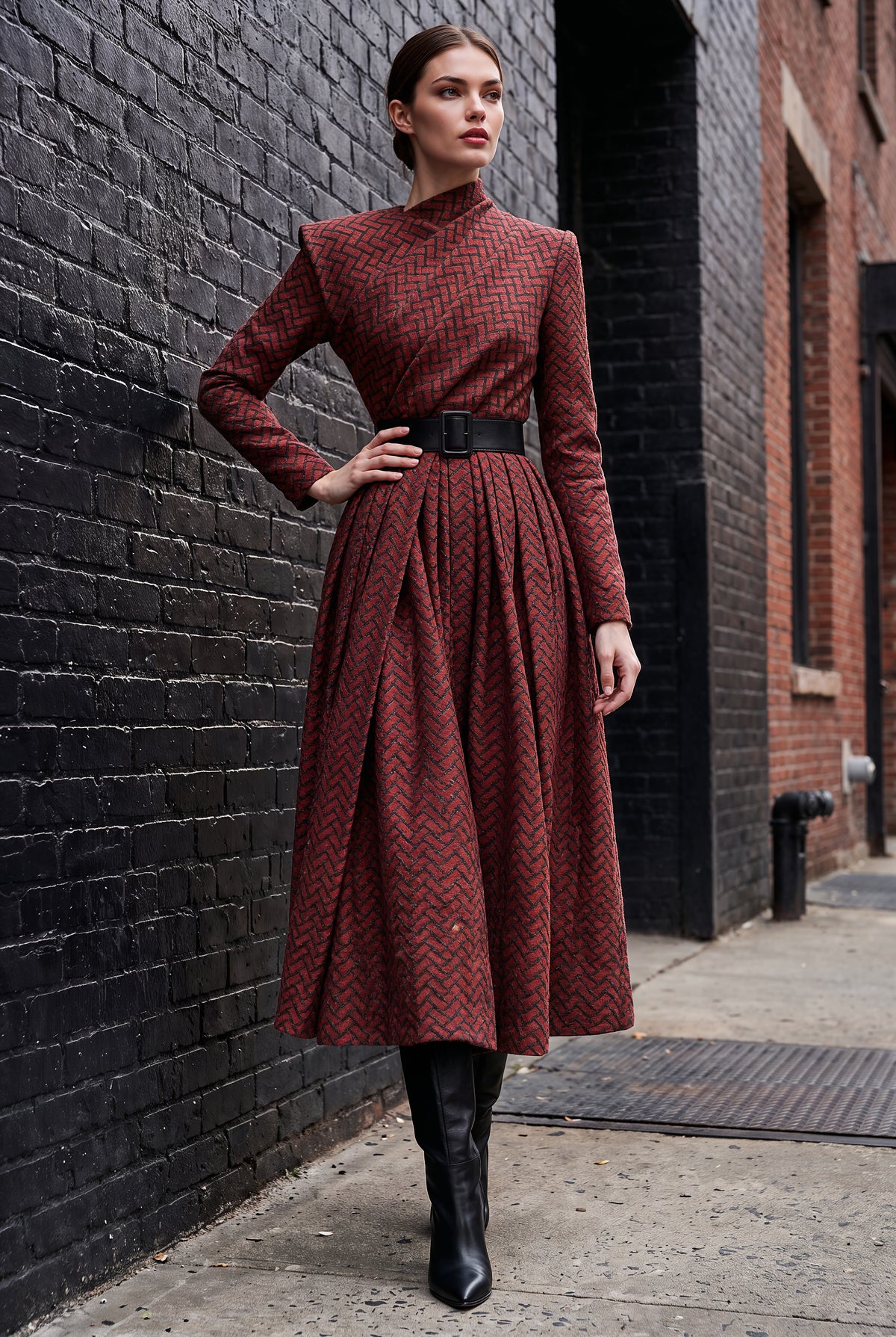A woman stands on a city sidewalk against a black brick wall, wearing a long-sleeve midi dress with a red and black chevron pattern, a black belt, and black knee-high boots.