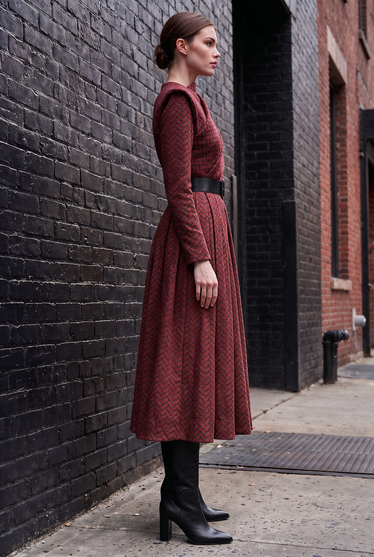 A woman stands in profile on a city sidewalk beside a black brick wall, wearing a long-sleeve midi dress with a textured chevron pattern in rust red and dark brown, a black belt, and black heeled ankle boots.
