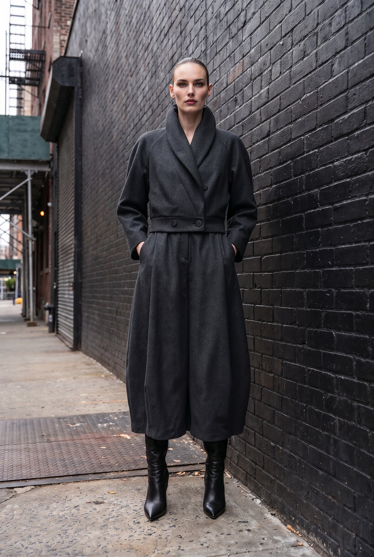 A model stands on a city sidewalk against a black brick wall, wearing a long black wrap-style coat with a belted waist and black ankle boots.