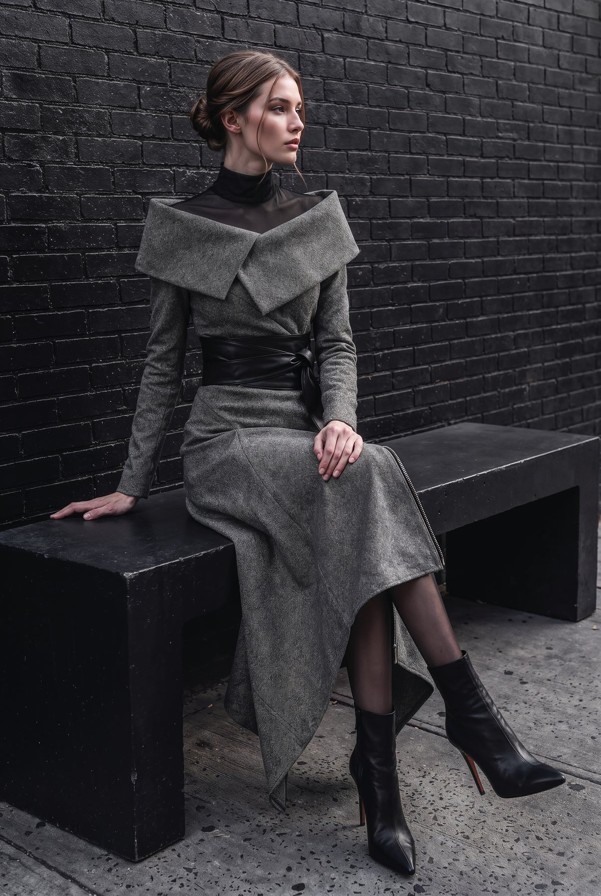 A model wearing a gray asymmetrical long-sleeve dress with a wide collar and black belt, sitting on a black bench against a dark brick wall.