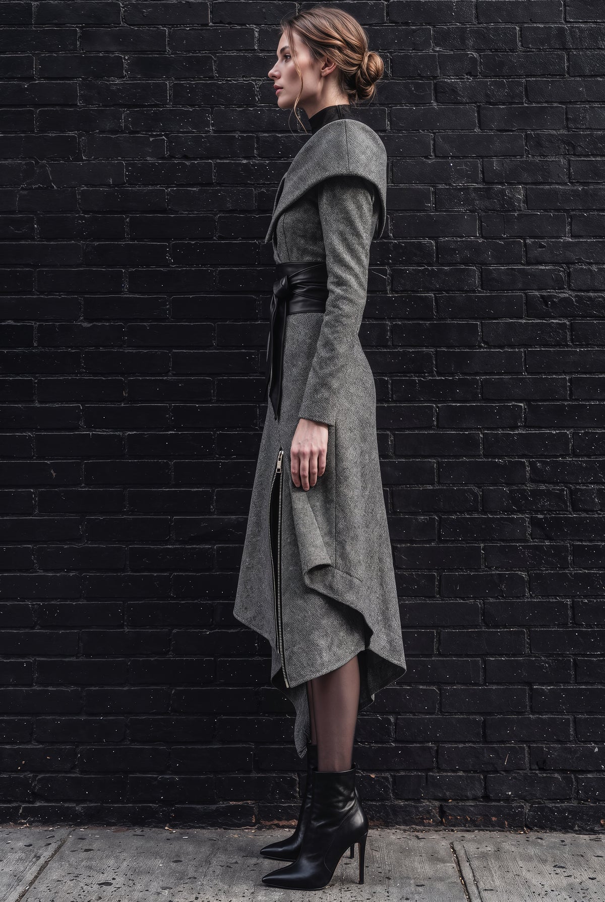 A woman stands in profile against a black brick wall wearing a long, draped gray wrap-style coat with a black belt, black tights, and black ankle boots.