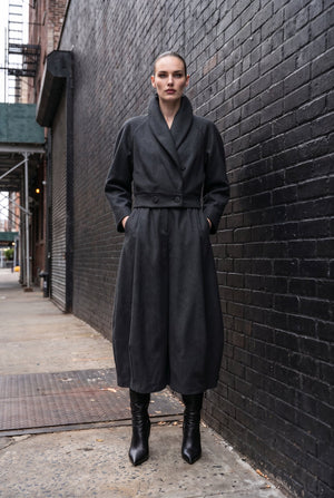 A model stands on a city sidewalk against a black brick wall, wearing a long black wrap-style coat with a belted waist and black ankle boots.