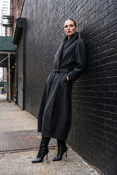 A woman wearing a long black wool coat with a belted waist and black knee-high heeled boots, leaning against a black brick wall on a city sidewalk.